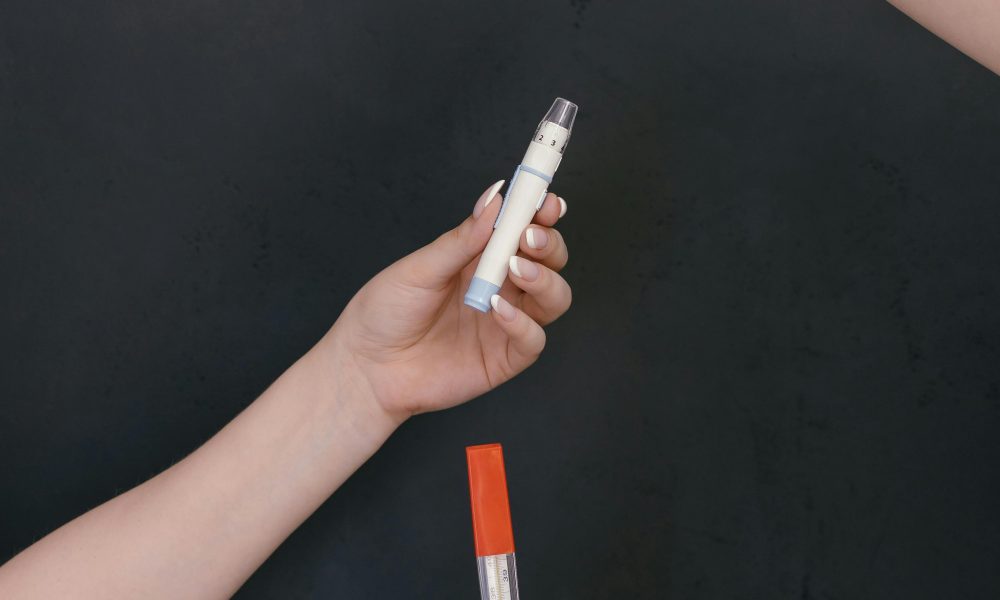 Three hands holding a cup, pen, and thermometer against a black backdrop, symbolizing healthcare.