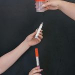 Three hands holding a cup, pen, and thermometer against a black backdrop, symbolizing healthcare.