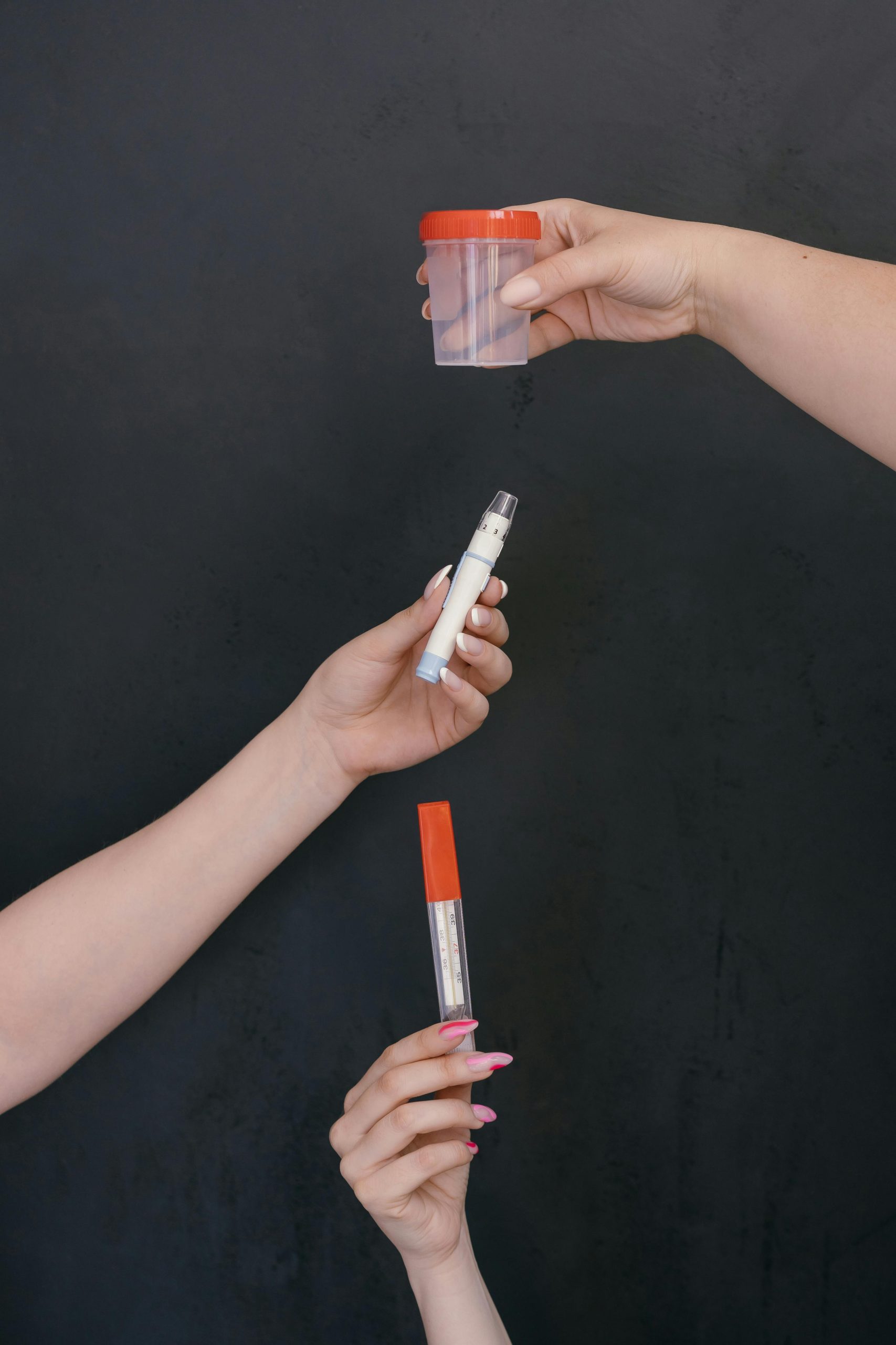 Three hands holding a cup, pen, and thermometer against a black backdrop, symbolizing healthcare.